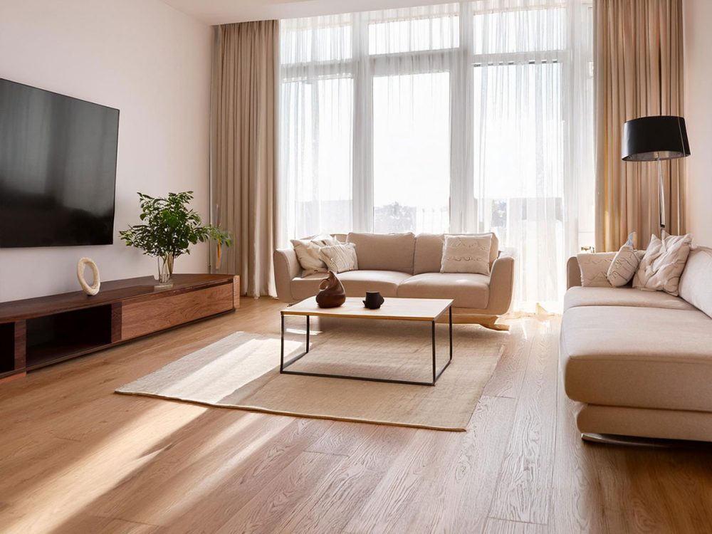 大きな窓から光が差し込むリビングに敷かれた無垢フローリングと、ベージュのソファが並ぶ落ち着いた空間。