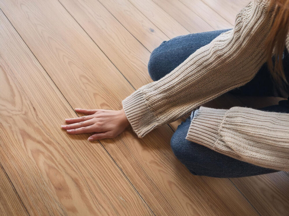 無垢フローリングに手を触れる女性。木目の質感や温もりを直接感じられる自然素材の床のイメージ。