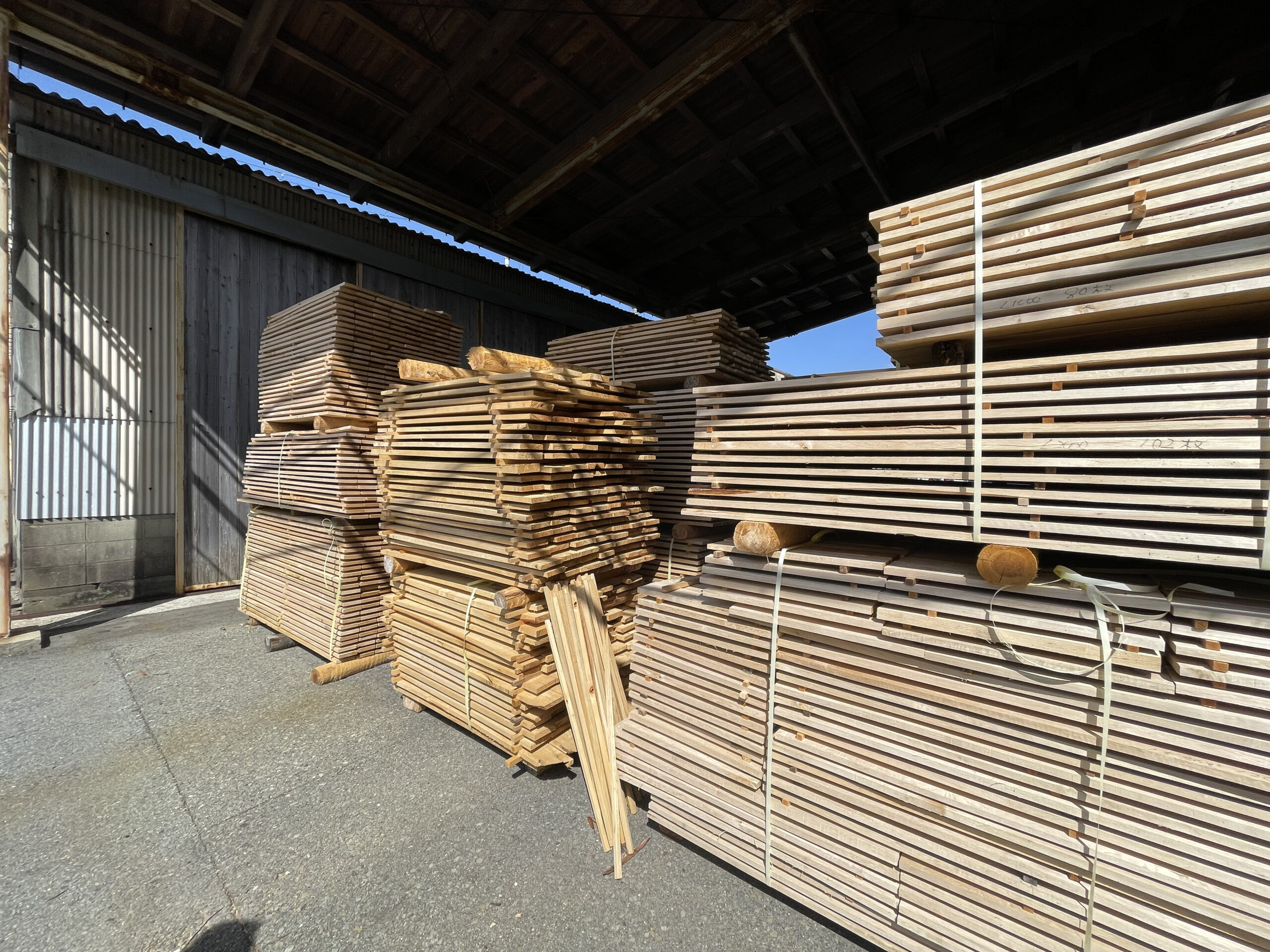 青空の下、屋外で桟積みされた無垢材が並ぶ様子。風通しの良い場所で自然乾燥が進んでいる。