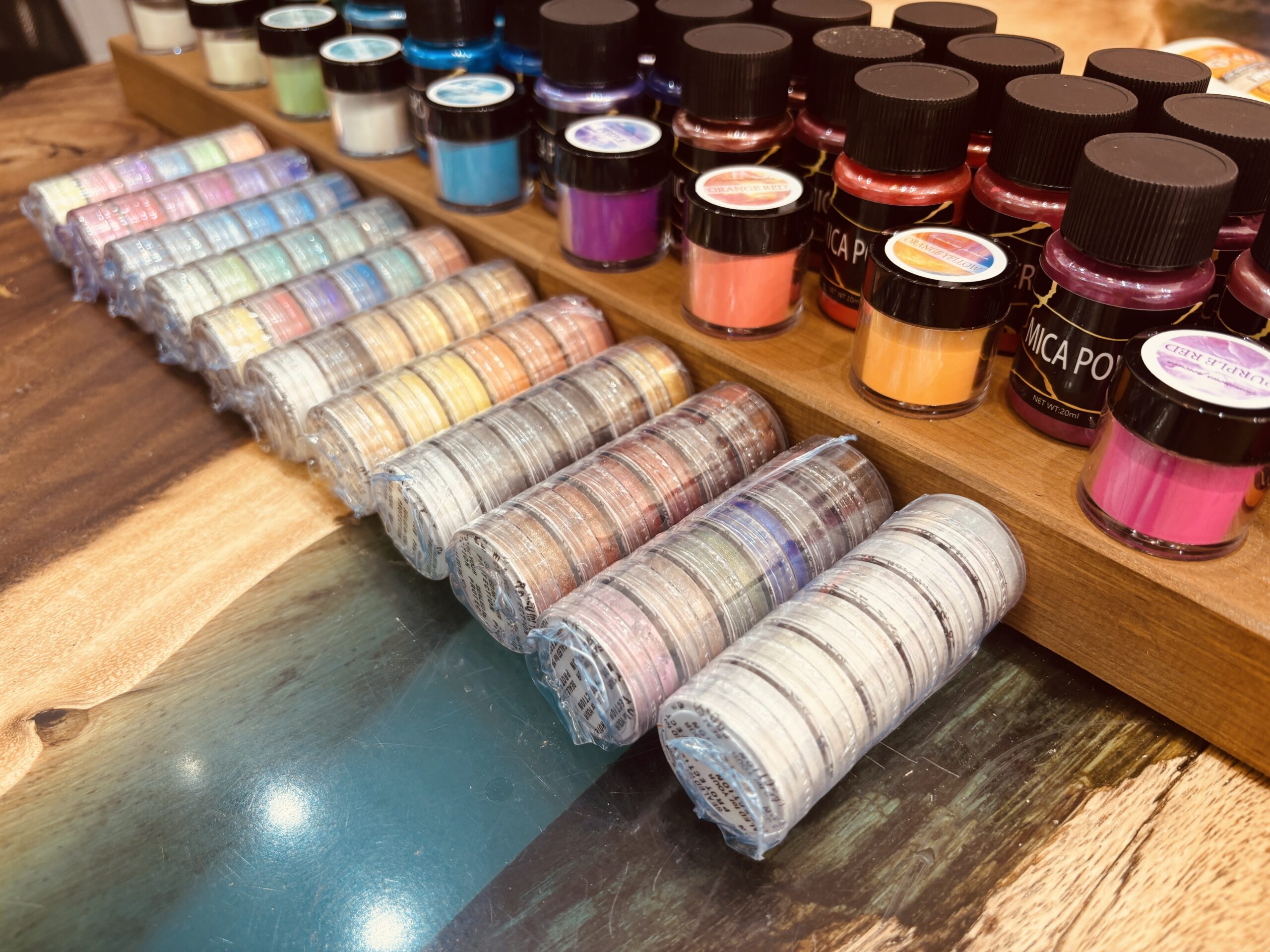 木目の上に並べられた多彩なマイカパウダー。下にはブルーのレジンが輝くレジンテーブルが見える。