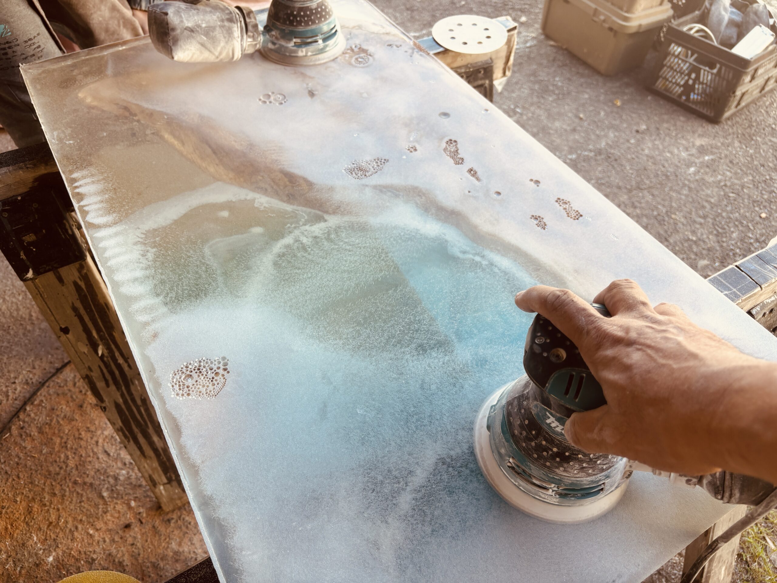 レジンテーブルの天板を電動サンダーで丁寧に研磨している様子。青みがかったレジン表面が白く曇り、加工途中であることがわかる。