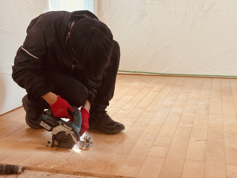 無垢フローリングの張り替え工事で、職人が丸ノコを使って既存の床材を丁寧に切り外している様子。床には木粉が広がり、慎重に作業を進めている姿が写っている。