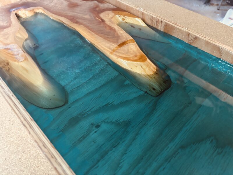 奈良県産カイヅカイブキを入江の形にくり抜き、ブルーグリーンのレジンを流し込んだレジンテーブルの天板。型枠の中で硬化途中の様子。