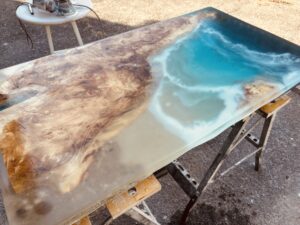 透明度の高いブルーグラデーションのレジンと、複雑な杢目を持つ木材が組み合わさったレジンテーブル天板を屋外で研磨している途中の様子。右側の海のようなブルーと白波の表現が際立つ。