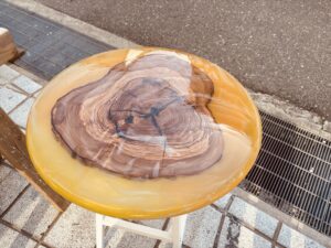 オリーブ材を中央に据え、オレンジ色のレジンで包み込んだ丸テーブル。自然光の下で、レジン内部の微細なラメがキラキラと輝き、磨いた部分と未研磨部分で透明度に差がある様子が分かる。