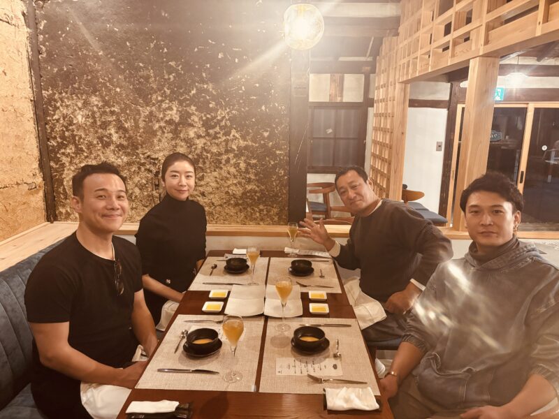 撮影後の打ち上げとして、落ち着いた雰囲気の洋食店「ケムリ」でテーブルを囲む4名のメンバー。温かい照明と木の interior が印象的な店内で、リラックスした表情で食事を楽しんでいる。