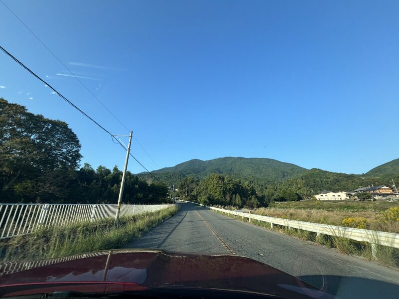 澄み切った青空と緑深い山々に囲まれた御所市の道路。車のフロント越しに見える、のどかな山あいのドライブ風景。