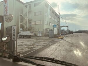 産廃処理施設の入口ゲートと建物。入口専用の看板、計量場、停車するトラックが見える朝の施設外観。