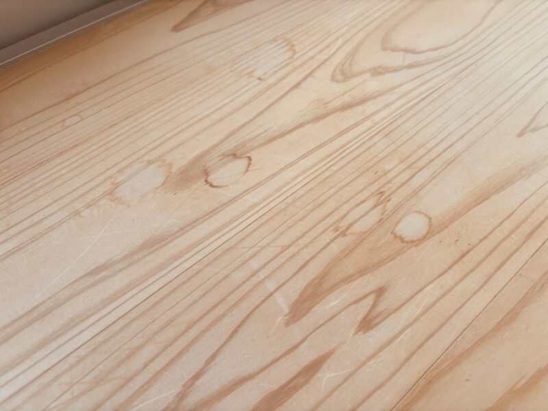 白濁した水じみと細かな傷が広がり、木目がくすんだ状態の杉フローリング。部分的に濃く染みが残っている。