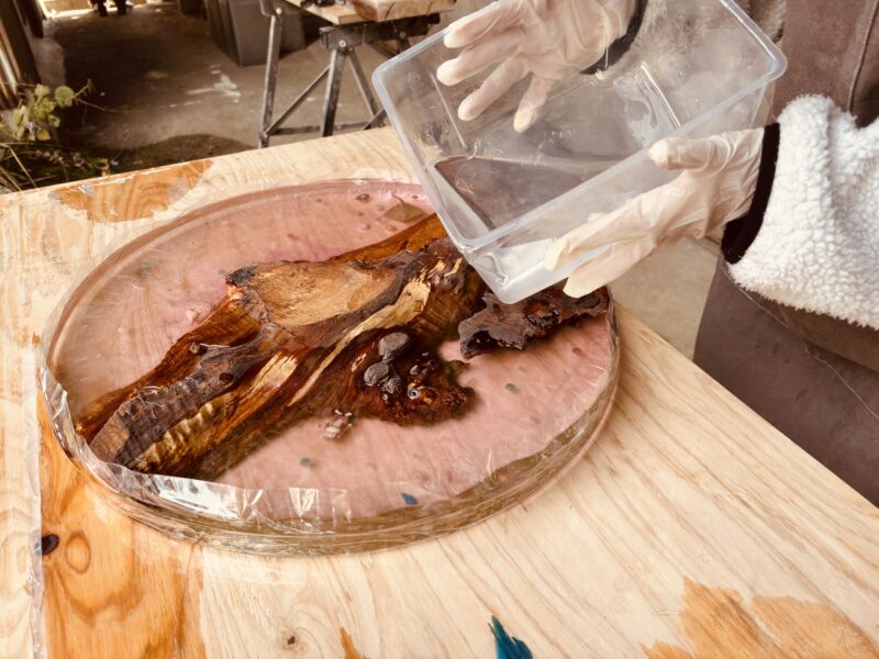 透明なレジンをラウンドテーブルの木材にゆっくりと注ぎ込む参加者さんの手元。木目がレジンに包まれ、新しい表情を見せ始めている瞬間。