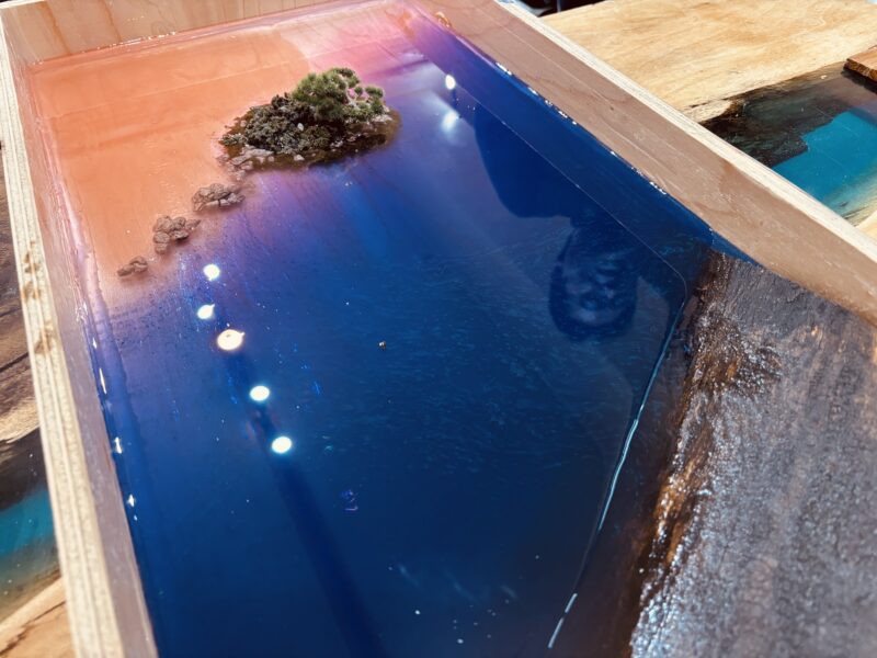 オレンジとネイビーの二層レジンが広がる中、義経岩をイメージした小島が浮かぶレジンテーブル制作のディテール写真。