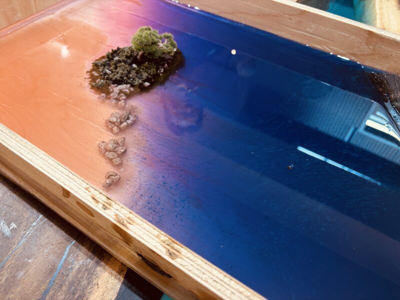 夕焼けの淡いピンクからネイビーの深海へと続くレジン層の上に、義経岩をイメージした島と岩が配置された制作途中のテーブル作品。