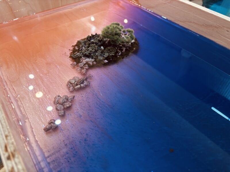 夕焼け色のオレンジから深いブルーへと滑らかに変化するレジンの海に、義経岩を模した小島と小石が並ぶ制作途中のレジンテーブル作品。