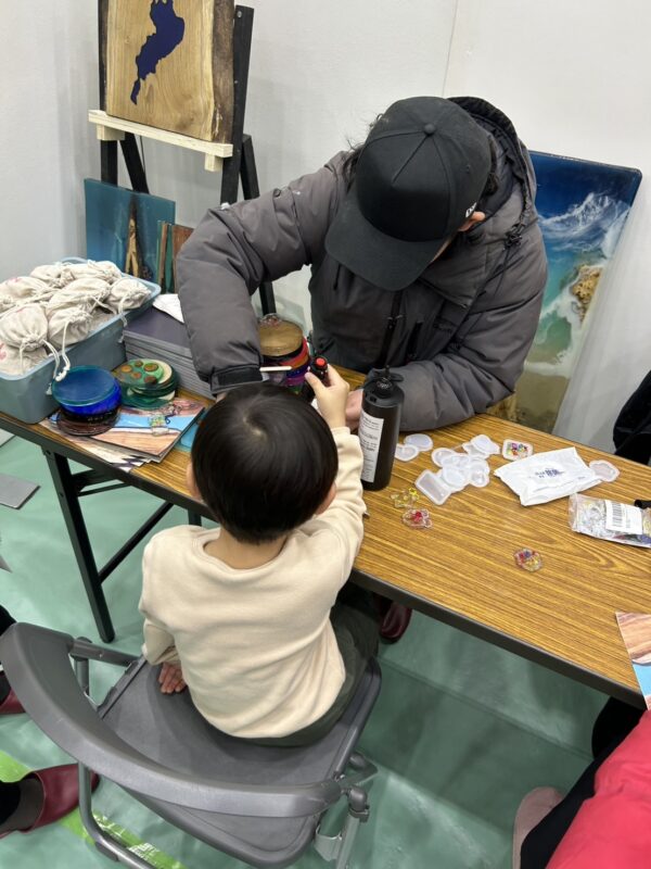 けんしんビジネスマッチングのエコロキアブースで、親子でレジンキーホルダー制作体験を楽しむ様子