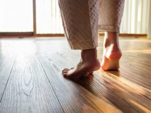 朝の光が差し込む無垢フローリングの上を素足で歩く女性の足元。木の温もりと心地よさを感じられる室内シーン。
