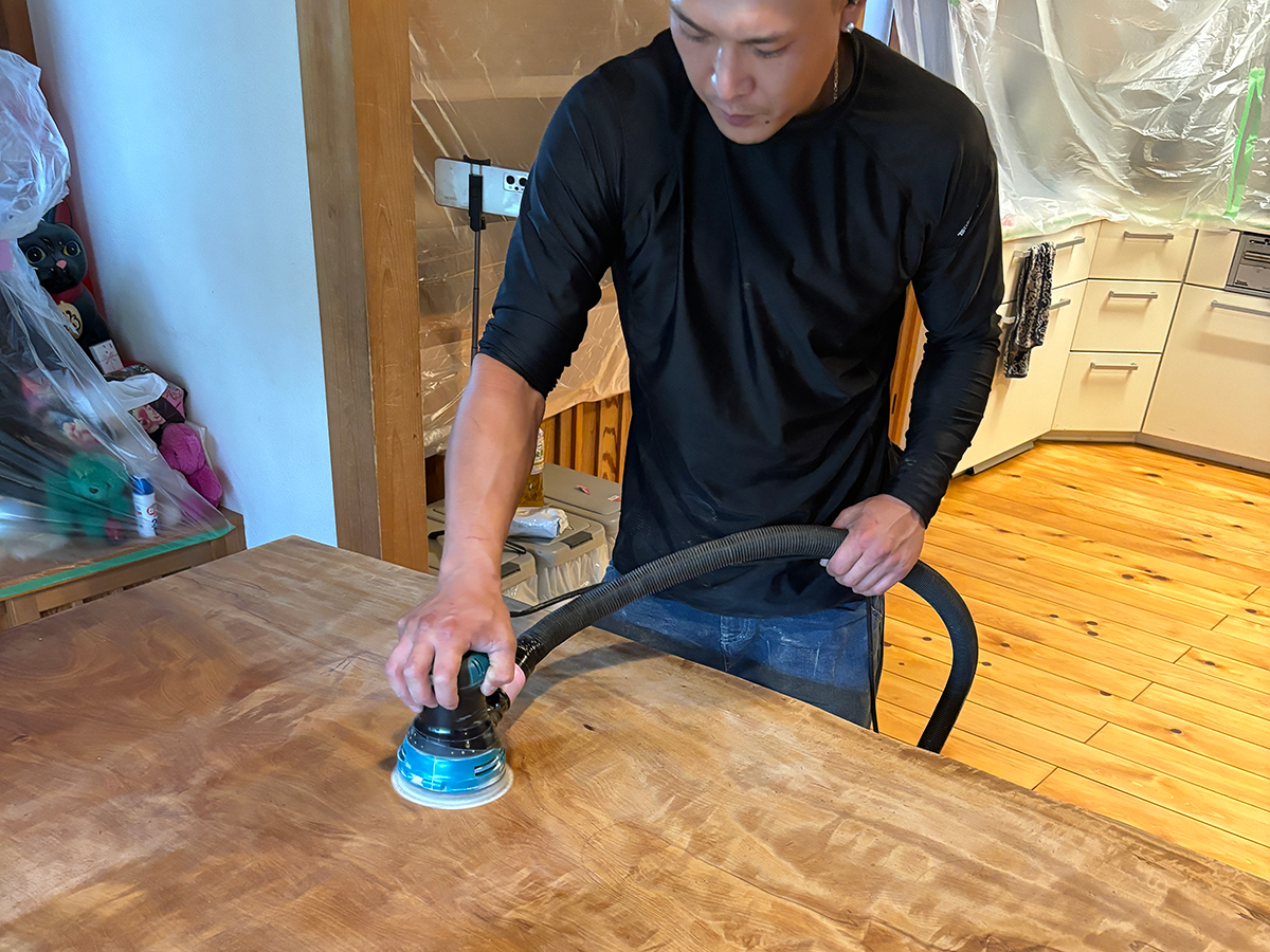 集塵機付きサンダーで栃の一枚板テーブルを研磨している様子。粉塵を抑えながら均一に表面を整えている。
